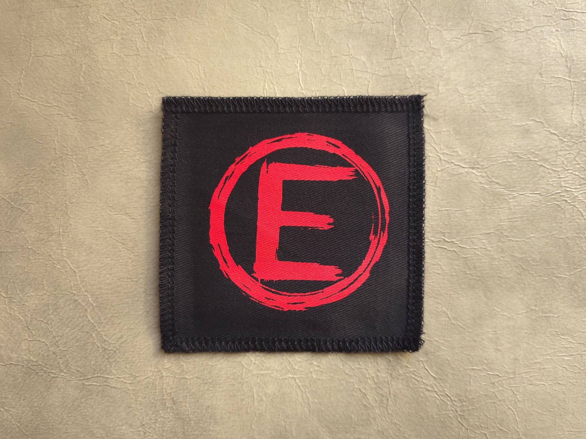 handmade screen printed Black patch with a red 'E' in the center on a beige background. equality symbol
